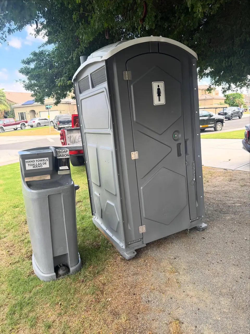 Gray standard porta potty with a handwashing station beside it on a lawn